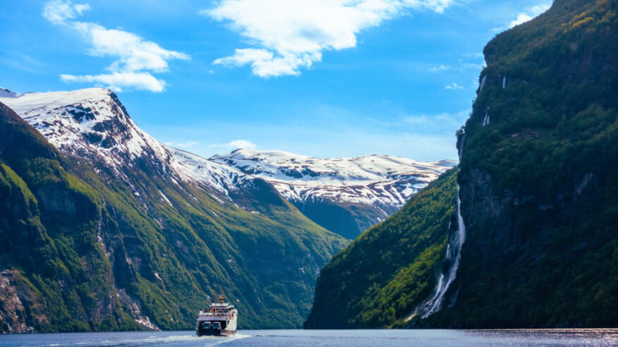 Cruising the majestic Geirangerfjord in the gorge inlet between steep snowcapped mountains lined with lush green trees, foliage and powerful waterfalls and flowing water from melted snow on a mostly blue sky with partial clouds