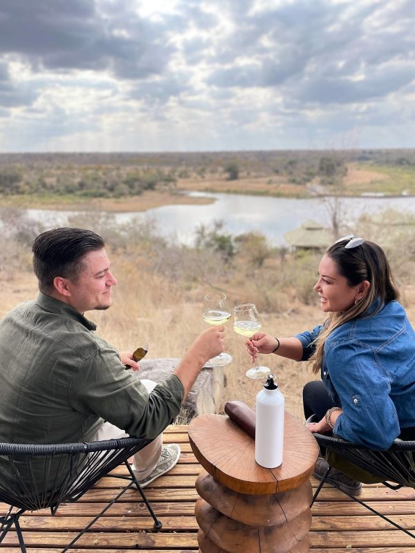 A man and a woman sitting on a deck overlooking a waterway and savannah in South Africa while toasting with wine glasses.